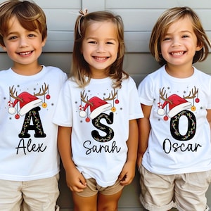 May include: Three children in white t-shirts with festive Christmas designs. Each shirt displays a letter, a Santa hat, reindeer antlers, and baubles. The names "Alex", "Sarah", and "Oscar" are printed below the letters.