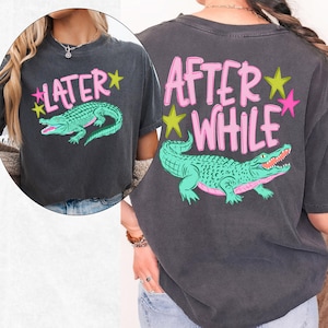May include: Dark gray t-shirt with a teal alligator graphic and the text "LATER" on the front and "AFTER WHILE" on the back, in pink lettering with yellow and pink star accents. The shirt has a vintage, worn look.
