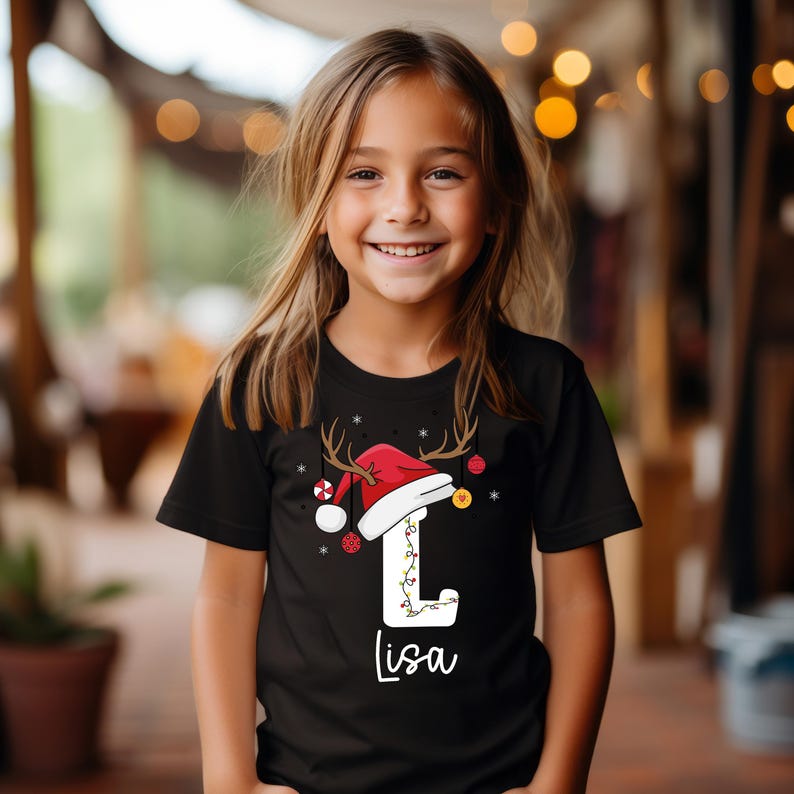 May include: Black t-shirt with a Christmas design. It features a red Santa hat with antlers, the letter "L" adorned with lights, and the name "Lisa" in cursive. The design is completed with festive baubles, perfect for the holiday season.