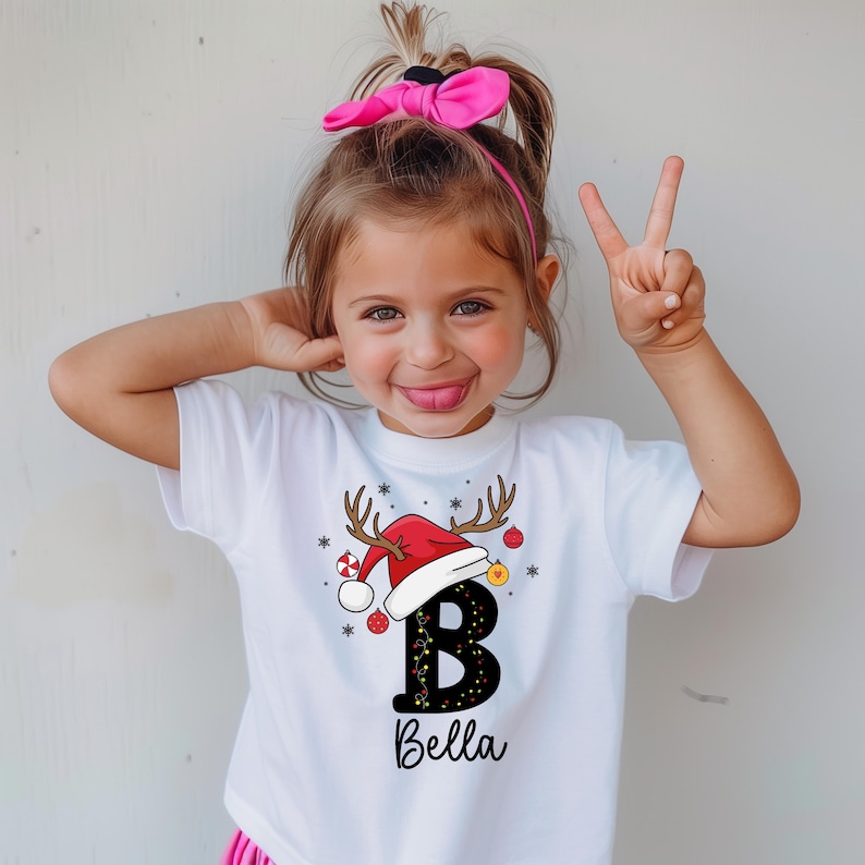 May include: White t-shirt featuring a Christmas design. The design includes a red Santa hat with antlers, the letter "B" decorated with lights, and the name "Bella" in a script font. The shirt is worn by a young child.