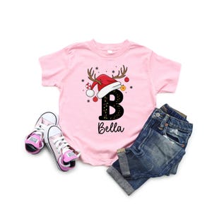 May include: A pink short-sleeved t-shirt with a Christmas design, featuring a red Santa hat with antlers and the letter "B" with the name "Bella" below. The shirt is paired with blue denim shorts and pink trainers.