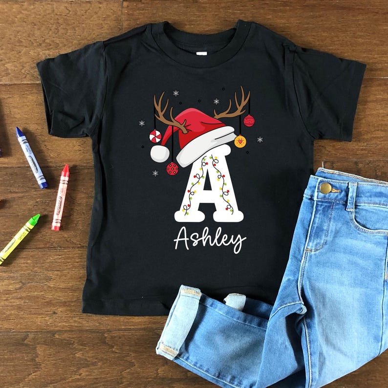 May include: Black t-shirt with a festive Christmas design. The design includes a red Santa hat with antlers, baubles, and the letter "A" adorned with lights. The name "Ashley" is written below. Blue jeans are next to the shirt.
