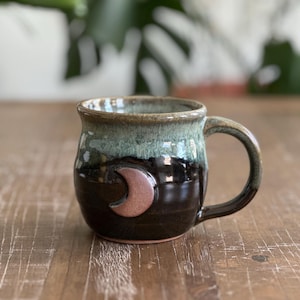 May include: A handcrafted ceramic mug with a crescent moon motif. The mug has a dark brown base, a green and black upper section, and a brown handle. The moon is a light brown colour. The mug is on a wooden surface.