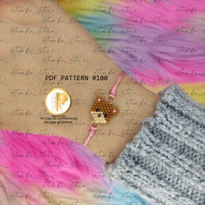 Op de afbeelding: Een kralenarmband met een bruin berenontwerp. De beer is gemaakt van kleine, kleurrijke kralen. De armband heeft een roze koord en goudkleurige metalen ringen. De tekst "PDF PATTERN #100" is zichtbaar.