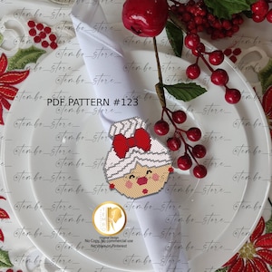 Puede incluir: Un servilletero navideño de cuentas con forma de mujer de jengibre con un lazo rojo. El servilletero está colocado sobre una servilleta blanca, con bayas rojas y una manzana roja en el fondo. El texto "PDF PATTERN #123" es visible.