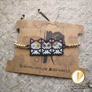 Puede incluir: Pulsera de cuentas con tres caras de gato en negro, blanco, rosa y dorado. La pulsera tiene cuentas doradas a cada lado. Se muestra en una etiqueta marrón con el texto "Tembo Store" y redes sociales.