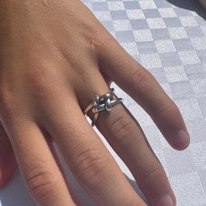 May include: A silver-colored ring with a knot design. The ring features a woven knot detail at the center, with a smooth, polished band. The ring is worn on a finger, showcasing its simple yet elegant design.