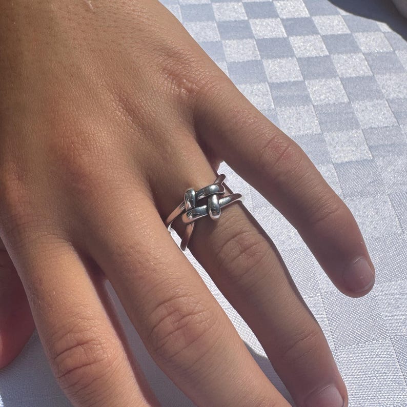 May include: A silver-colored ring with a knot design. The ring features a woven knot detail at the center, with a smooth, polished band. The ring is worn on a finger, showcasing its simple yet elegant design.