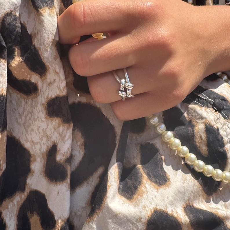 May include: A silver-colored ring with a knot design. The ring is worn on a finger, with a leopard print fabric in the background. A strand of pearls is also visible.