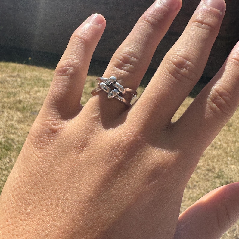 May include: A silver-colored ring with a woven design. The ring features a series of interconnected loops, creating a textured and intricate pattern. The ring is worn on a finger, showcasing its design and craftsmanship.