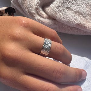 Puede incluir: Un anillo de plata con una superficie texturizada. El anillo es ancho y tiene un diseño martillado u orgánico. La joya se lleva en un dedo.