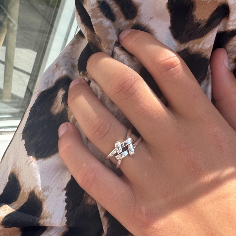 May include: A silver-colored ring with a woven knot design. The ring is worn on a finger, with a leopard print fabric in the background. The ring's design features intersecting bars, creating a unique and stylish accessory.