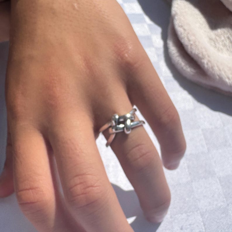 May include: A silver-colored ring with a knot design. The ring is worn on a finger. The ring's design features a series of interconnected loops, creating a textured and eye-catching appearance. The ring is made of a shiny metal.