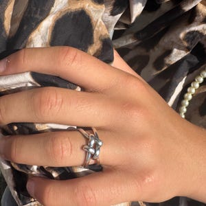 May include: A silver-colored ring in a knot design. The ring is on a hand with a leopard print fabric in the background. A strand of pearls is also visible.