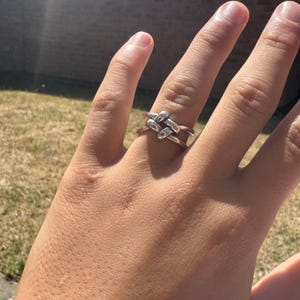 May include: A silver-colored ring with a woven design. The ring features a series of interlocking, rectangular shapes, creating a textured and geometric pattern. The ring is worn on a finger, with the sunlight reflecting off its surface.