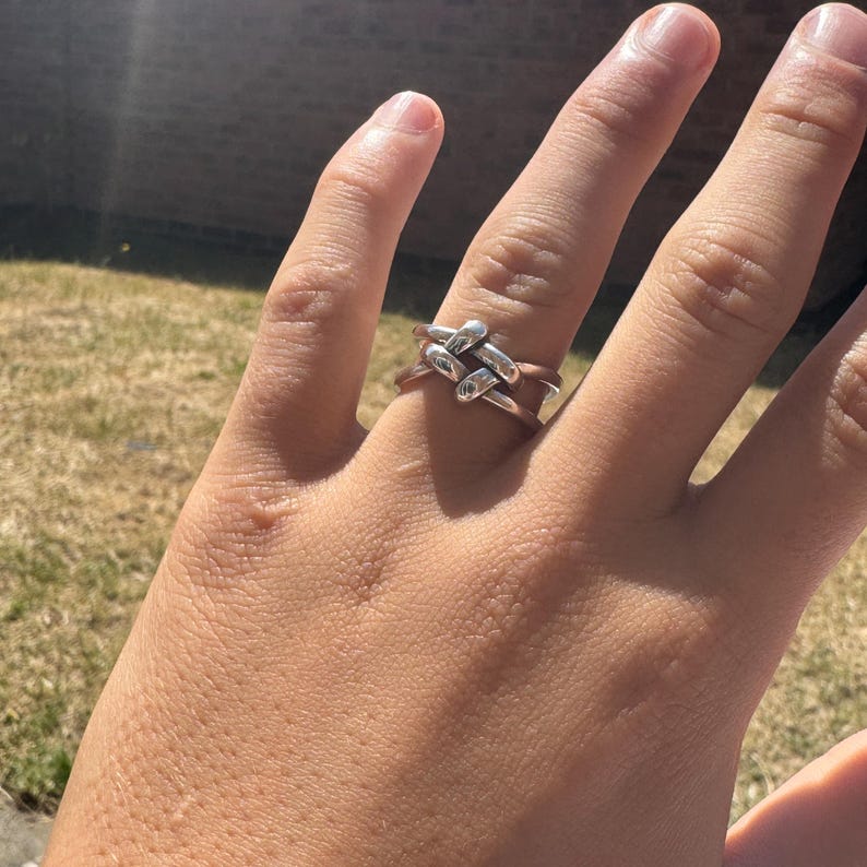 May include: A silver-colored ring with a woven design. The ring features a series of interlocking, rectangular shapes, creating a textured and geometric pattern. The ring is worn on a finger, with the sunlight reflecting off its surface.