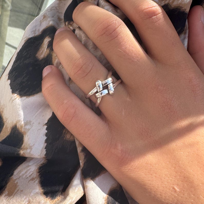 May include: A silver-colored ring with a unique knot design. The ring features a series of intersecting bars, creating a textured and eye-catching appearance. The ring is worn on a finger, showcasing its intricate details and polished finish.