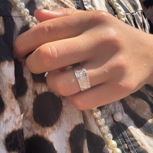 Op de afbeelding: Een zilveren ring met een getextureerd oppervlak wordt aan een vinger gedragen. De ring heeft een bandvormig ontwerp en lijkt van metaal te zijn. De achtergrond omvat een patroonstof en een parelsnoer.