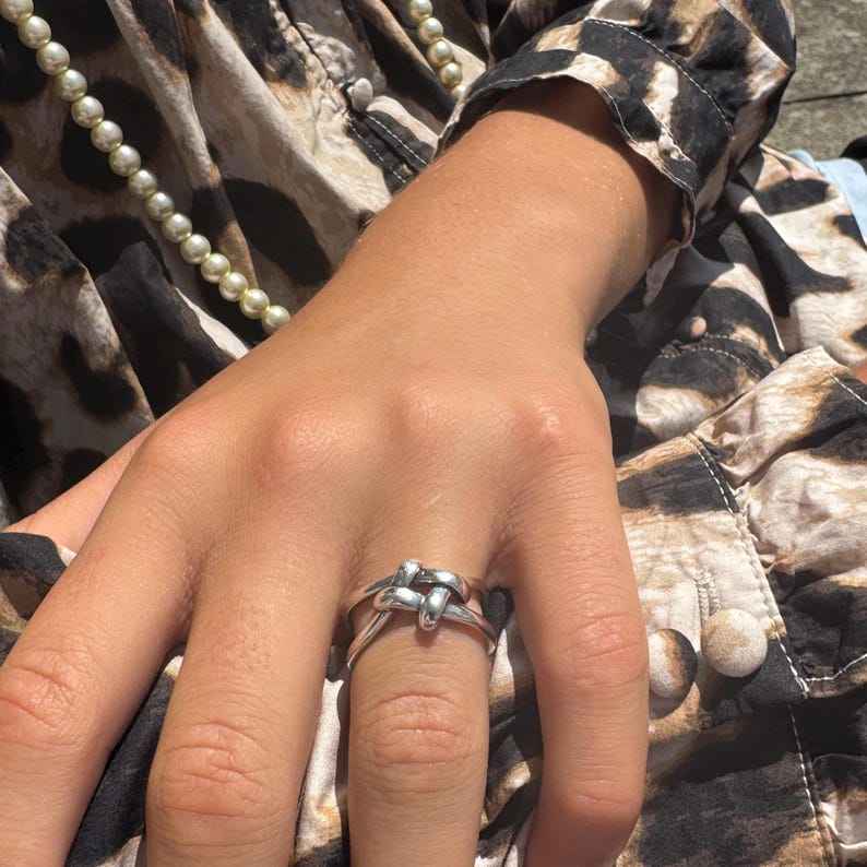 May include: A silver-colored ring with a knot design. The ring is worn on a finger, with a patterned fabric and a pearl necklace in the background. The ring's knot design is the focal point, showcasing a simple yet elegant style.