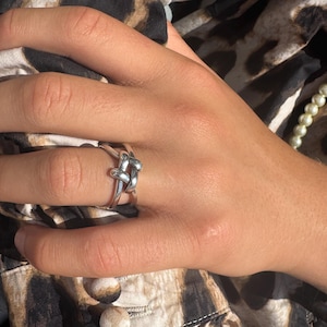 May include: A silver-colored ring with a knot design. The ring is on a hand with a leopard print background. The ring's knot is the focal point, with the band appearing to be two intertwined loops.