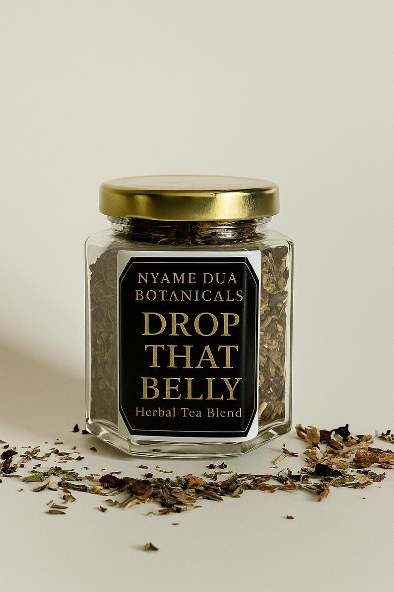 May include: A clear glass jar with a gold lid, filled with herbal tea blend. The label reads "NYAME DUA BOTANICALS DROP THAT BELLY Herbal Tea Blend." Loose tea leaves are scattered around the jar.