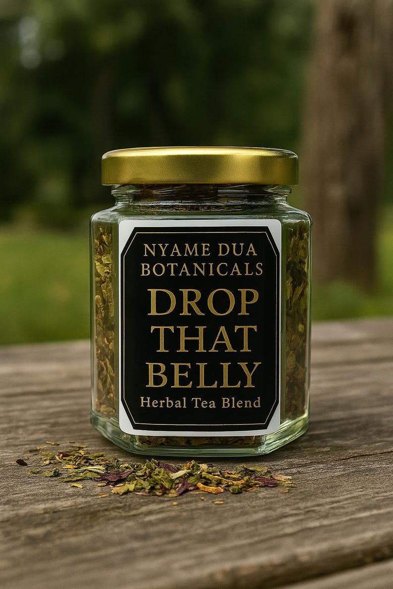 May include: A clear glass jar with a gold lid, filled with dried green herbs. The label reads "NYAME DUA BOTANICALS DROP THAT BELLY Herbal Tea Blend" in gold lettering on a black and white background. The jar sits on a wooden surface.