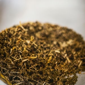 May include: A close-up shot of dried herbs in a clear glass container. The herbs are a mix of green, brown, and yellow, with various textures. The image is well-lit, showing the details of the herbs.