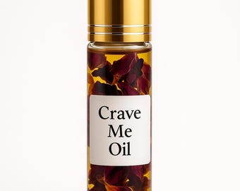 May include: A clear glass roll-on bottle with a gold cap, filled with a golden liquid and red petals. The label reads "Crave Me Oil". The bottle is set against a white background.