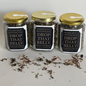 May include: Three glass jars with gold lids filled with herbal tea blend. Each jar has a black and white label that reads "NYAME DUA BOTANICALS DROP THAT BELLY Herbal Tea Blend". Loose tea leaves are scattered around the jars.