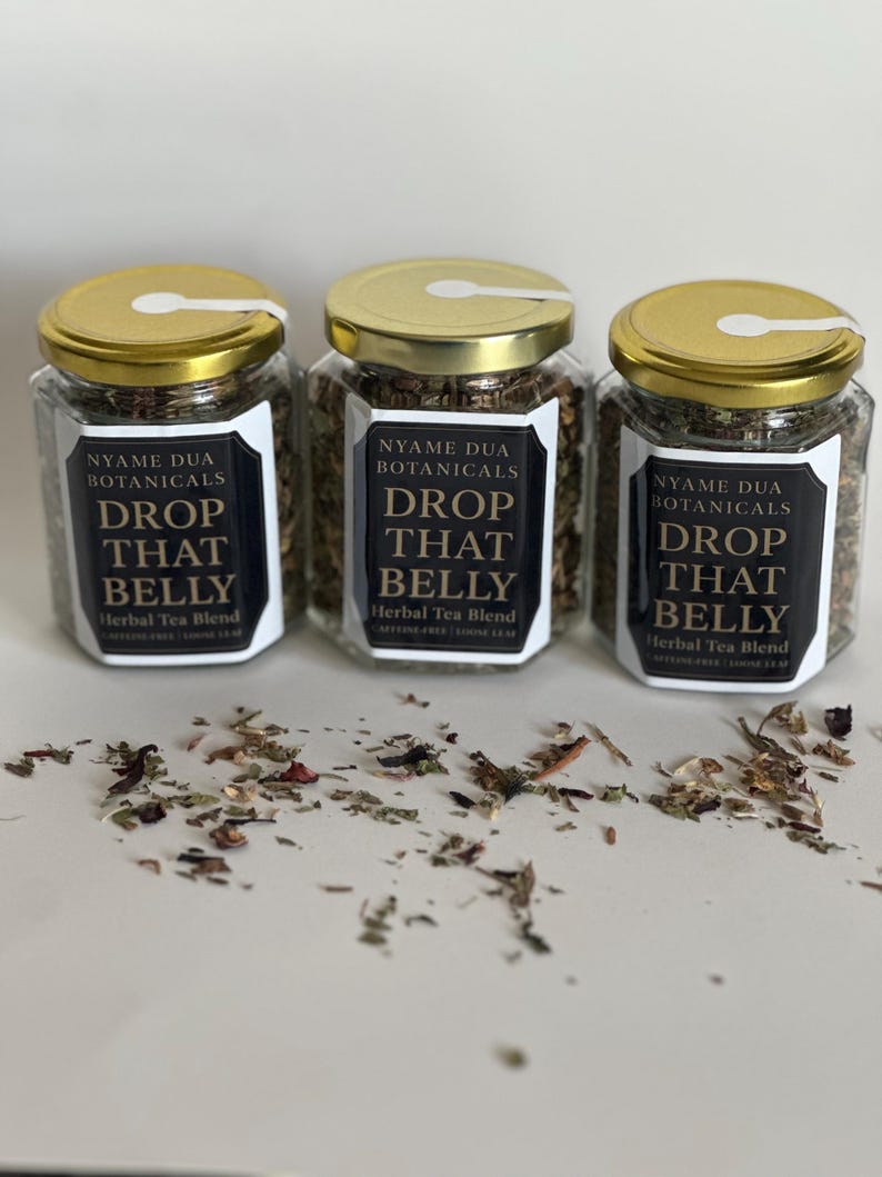 May include: Three glass jars with gold lids filled with herbal tea blend. Each jar has a black and white label that reads "NYAME DUA BOTANICALS DROP THAT BELLY Herbal Tea Blend". Loose tea leaves are scattered around the jars.