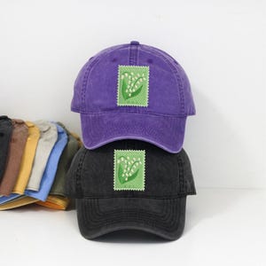 Könnte beinhalten: Zwei Baseballkappen, eine lila und eine schwarze, übereinander gestapelt. Jede Kappe hat ein grünes Briefmarken-Design mit weißen Blumen. Im Hintergrund sind weitere farbige Kappen zu sehen. Ein modisches Accessoire.