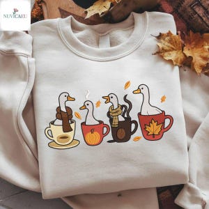 Può includere: Felpa color crema con un design stravagante di quattro anatre in tazze da caffè. Ogni anatra è raffigurata con una sciarpa e un design di tazza diversi, tra cui una zucca e una foglia d'acero. Foglie d'autunno sparse.