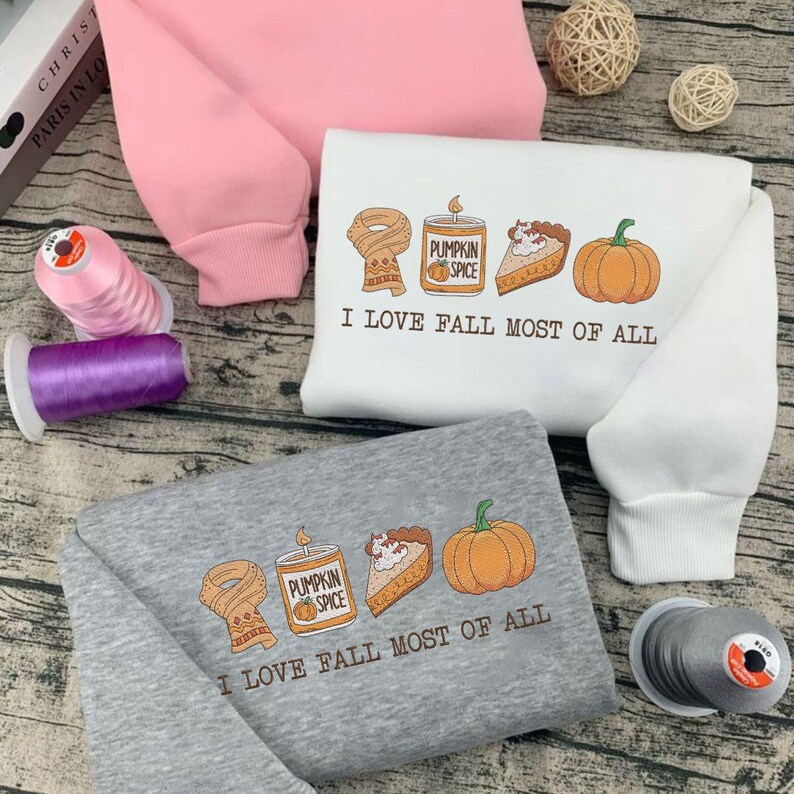 May include: Three sweatshirts in pink, white, and gray, each featuring a fall-themed graphic. The design includes a scarf, pumpkin spice candle, pie slice, and pumpkin, with the text "I LOVE FALL MOST OF ALL".