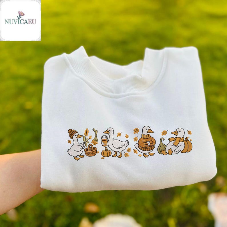 May include: White sweatshirt featuring an embroidered design of four ducks in autumn attire. The ducks are depicted with a hat, a coffee cup, a sweater, and a scarf, along with pumpkins and leaves. The brand name "NUVICAEU" is in the top left corner.