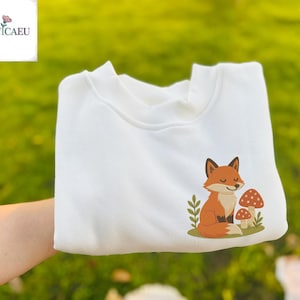 Peut inclure: Un sweat-shirt blanc avec un motif brodé d'un renard de dessin animé et de champignons. Le renard est orange et blanc avec un nez noir et les yeux fermés. Les champignons sont rouges et blancs.