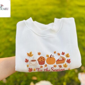 May include: White sweatshirt with an embroidered fall-themed design. The design includes a coffee cup, muffin, pumpkin, and pie, surrounded by autumn leaves. The text "Tis the Season" is below the design.