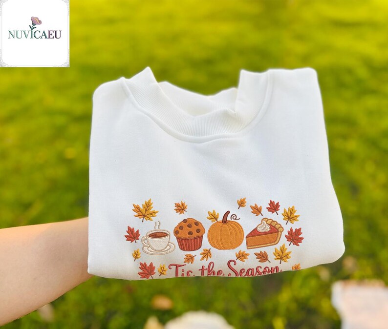 May include: White sweatshirt with an embroidered fall-themed design. The design includes a coffee cup, muffin, pumpkin, and pie, surrounded by autumn leaves. The text "Tis the Season" is below the design.