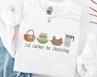 May include: White crewneck sweatshirt with embroidered images of a basket, bowls, a chicken, and a pitcher. The text "I'd rather be thrifting" is embroidered below the images. A silver necklace with a small pendant is also visible.