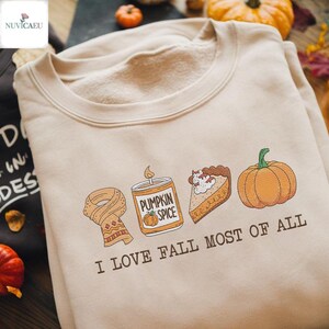 May include: Beige sweatshirt with a crew neck and the phrase "I LOVE FALL MOST OF ALL." The graphic includes a scarf, pumpkin spice candle, slice of pie, and a pumpkin.