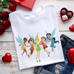 Könnte beinhalten: Weißes T-Shirt mit einem gestickten Design von sechs Feen mit bunten Flügeln und Kleidern. Die Feen sind in Gelb-, Orange-, Grün- und Rottönen gehalten. Das Shirt liegt auf einer Holzoberfläche mit Rosen, Pralinen und Jeansshorts.