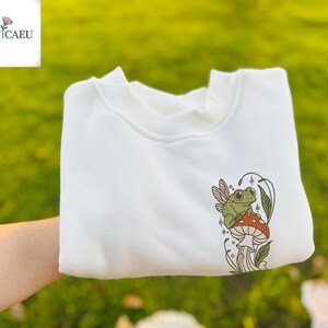 Könnte beinhalten: Weißes Sweatshirt mit einem skurrilen Design einer grünen Frosch mit Feenflügeln auf einem roten Pilz. Das Design umfasst Sterne und Blätter. Das Sweatshirt hat einen Rundhalsausschnitt.