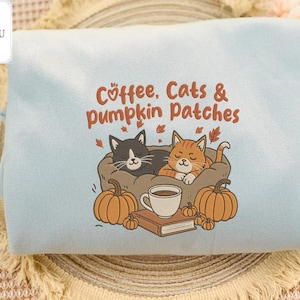 Peut inclure: Sweat-shirt bleu clair avec l'inscription "Coffee, Cats & Pumpkin Patches". Le motif représente un chat noir et un chat roux dans un lit, des citrouilles, une tasse de café et un livre. Un article sur le thème de l'automne.