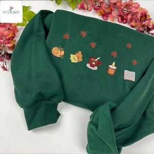 May include: A dark green sweatshirt with embroidered autumn-themed designs. The design includes a pumpkin, leaves, a cup of tea, a candle, and a book. The sweatshirt has long sleeves and a crew neck.