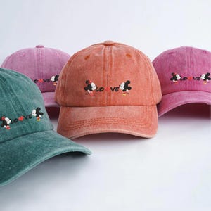 Op de afbeelding: Verschillende baseballpetten in kleuren als teal, roze en oranje. Elke pet heeft een geborduurd ontwerp van Mickey en Minnie Mouse met het woord "LOVE". De petten hebben een vintage uitstraling.