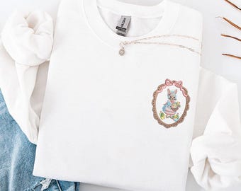 May include: White crewneck sweatshirt with an embroidered design. The design features a bunny in a dress holding a basket of eggs, framed in an oval with a pink bow. A silver necklace with a small pendant is also visible.