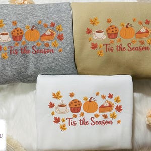 Könnte beinhalten: Drei Sweatshirts in Grau, Beige und Weiß, jeweils mit einem herbstlichen Design bestickt. Das Design umfasst eine Tasse Kaffee, einen Muffin, einen Kürbis, ein Stück Kuchen und Herbstblätter. Der Text "Tis the Season" ist ebenfalls gestickt.