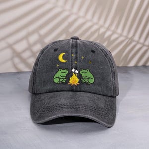 Può includere: Cappellino da baseball grigio scuro con un design ricamato. Due rane verdi arrostiscono marshmallow su un falò, con una luna crescente gialla e stelle sopra. Il cappellino ha una visiera curva e una chiusura con cinturino in tessuto.