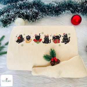 Könnte beinhalten: Cremefarbenes Kissen und Nackenrolle mit gesticktem Design von schwarzen Katzen in festlicher Kleidung. Die Katzen tragen Weihnachtsmützen, Rentiergeweihe und Lichterketten. Das Kissen ist mit Schneeflocken verziert.