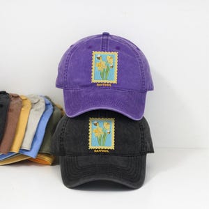 May include: Two baseball caps, one purple and one black, stacked on top of each other. Each cap features a daffodil stamp design with the word "DAFFODIL" below the image. The caps are made of a soft, worn material.