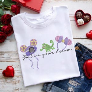 Könnte beinhalten: Weißes T-Shirt mit gestickten Designs einer lila-gelben Sonne, einem grünen Frosch und einer lila Schleife. Der Satz "Go live your dream" ist in schwarzer Schrift geschrieben. Das Shirt wird mit Jeansshorts kombiniert.